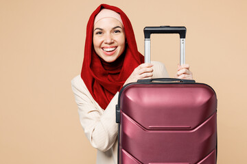 Wall Mural - Traveler happy Asian Muslim woman wears red abaya hijab hold suitcase bag isolated on plain beige background Tourist travel abroad in free spare time rest getaway Air flight trip journey, UAE concept