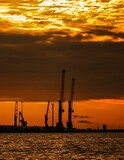 Fototapeta Most - Vertical shot of mining equipment and cranes on the shore of an industrial area at sunset
