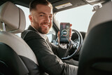 Fototapeta  - Monitoring charging process, app in smartphone. Man in black suit is sitting in the car, driving