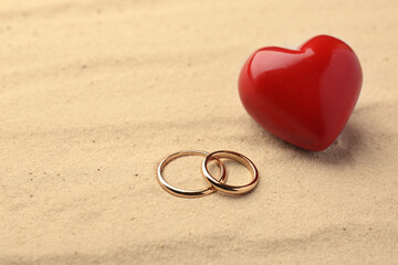 Honeymoon concept. Two golden rings and red wooden heart on sand