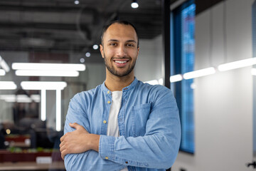 Portrait of a young hispanic male programmer and developer standing in the office, crossing his arms over his chest and looking at the camera with a smile.
