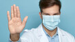 A man in a white lab coat is wearing a blue shirt and a blue mask. He is holding his hand up in a 