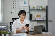 © apichat - Cheerful businesswoman in white shirt using smartphone with laptop and documents on her office desk.