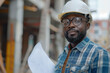 African American man in a hard hat and glasses is a builder with drawings, an engineer or an architect. He supervises the work and ensures compliance with safety protocols