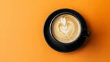 Top view of a black coffee cup with a latte heart design on the foam against an orange background.