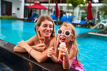 Cheerful young girl with sunglasses enjoys ice cream beside smiling mom in a pool. Relaxing family moment during sunny holiday, bonding over sweet treats, water in background, summer vibe.