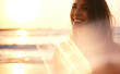 A smiling woman enjoys the golden light on a beach as the sun sets, with a soft focus and lens flare