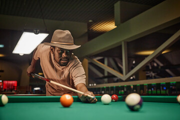 Wall Mural - Portrait of Black young man playing billiards and wearing hat while hitting ball with cue stick in low lighting copy space