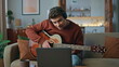 Talented guy playing guitar wearing headphones at couch. Calm man looking laptop