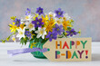 Bouquet of spring blue, purple, white and yellow flowers in a vase on the table and a card with the text happy birthday.
