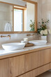 A closeup of two white ceramic basins on top, set in an elegant oak wooden bathroom cabinet with brass faucet and warm beige tiles, complemented in the style of soft natural light from a large window