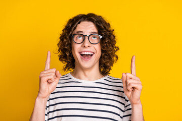 Sticker - Photo of handsome funny teenage guy in striped t shirt pointing fingers up promoting best offers isolated on yellow color background