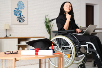 Poster - Beautiful female graduate student in wheelchair with graduation cap and diploma on table at home