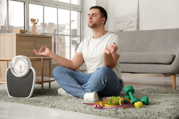 Poster - Young man with scales, dumbbells and food meditating at home. Weight loss concept