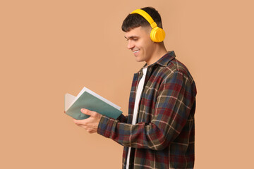 Poster - Handsome young happy man in headphones with book on beige background. Concept of audiobook