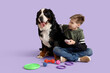 Little boy with pet toys and Bernese mountain dog sitting on lilac background