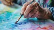 Elderly person's hand holding paintbrush, applying vibrant watercolor paints to canvas, showcasing creativity and artistry