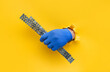 A left man's hand in a blue knitted glove holds a U-shaped fastening profile for drywall. Torn hole in yellow paper. The concept of a worker, a labor migrant, a master of his craft. Copy space.
