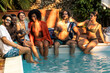 A diverse group of friends shares laughter and colorful drinks on a sunlit poolside - young people leisure and connection in summer setting.