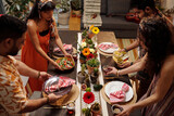 Fototapeta  - Two young couples in national attire putting drinks, homemade food and plates with napkins on festive table while serving it for dinner
