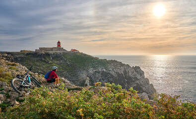 Wall Mural - happy active senior woman cycling during moody golden hour near sunset at the the rock cliffs and lighthouse of Cabo Sao Vicente, the south-western spit of Europe at the atlantic coast of Portugal