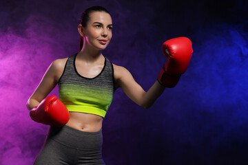 Sticker - Portrait of beautiful woman wearing boxing gloves training in color lights and smoke on black background