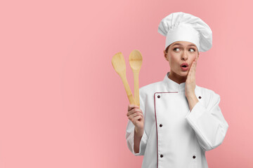 Wall Mural - Surprised chef in uniform holding wooden spatula and spoon on pink background, space for text