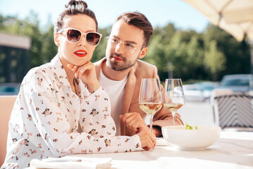 Wall Mural - Smiling beautiful woman and her handsome boyfriend. Happy cheerful family. Couple cheering with glasses of white wine at their date in restaurant. They drinking alcohol at veranda cafe in the street