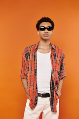 Wall Mural - trendy good looking african american man in vibrant attire with sunglasses on orange backdrop