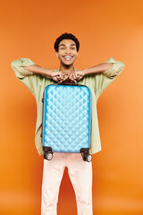 Wall Mural - cheerful handsome african american man in casual outfit holding blue suitcase and smiling at camera