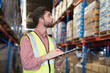 © amorn - Male warehouse worker holding clipboard during working in storage warehouse. Warehouse worker inspecting quality of products on shelf pallet and checking stock inventory
