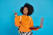 An African American college girl in casual attire holding a laptop, pointing to the side in a studio with a blue backdrop.