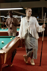 Wall Mural - Vertical full length portrait of bald young woman standing by pool table in nightclub and holding cue stick