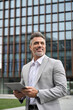 © insta_photos - Happy business man investor of middle age in suit holding tablet, mature professional businessman leader using tab looking away standing in busy office street thinking of investing in future. Vertical