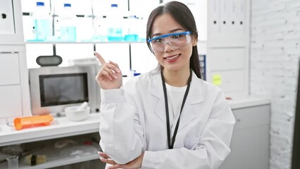 Sticker - Cheerful young chinese woman scientist confidently presenting with a radiant smile and pointing finger to the side in the lab