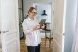 A contented woman with glasses uses a tablet, standing by a door in a stylish apartment.