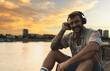 Young hipster millennial man sitting on riverside looking at city listening music on wireless headphones enjoying moment, life and sunset. Happy independent male enjoy freedom and life concept