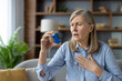 © Liubomir - An elderly woman experiences an asthma attack and uses a blue inhaler. She looks concerned while sitting in a modern living room.