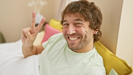 Sticker - Cheerful young caucasian man in pyjamas confidently shows number two, pointing up with two fingers from his cozy bedroom.