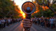 Hand holding a megaphone against a backdrop of a gathered crowd at sunset. Scene suggests a public event or protest.