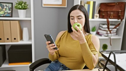 Wall Mural - Hispanic woman in office eating apple while browsing smartphone, portraying a casual work break.