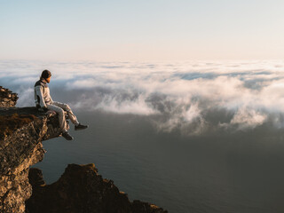 Wall Mural - man relaxing alone on the edge of mountain cliff in norway above clouds and sea, traveler hiking out