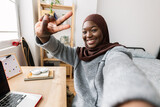 Fototapeta  - Cheerful young black african woman in muslim headscarf taking selfie portrait with phone at studio room.
