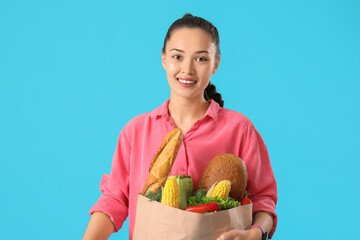 Poster - Beautiful young Asian woman holding paper bag with fresh products on blue background