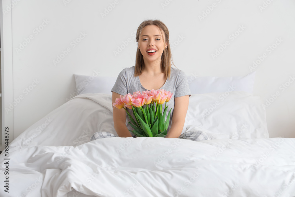 Happy young woman sitting on comfortable bed with bouquet of tulip flowers in bedroom