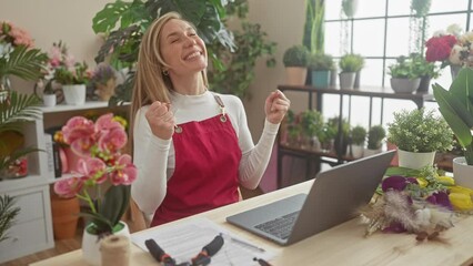 Poster - Joyful young blonde florist woman sitting at flower shop table, victoriously screaming in happy excitement celebrating win, gesturing success!