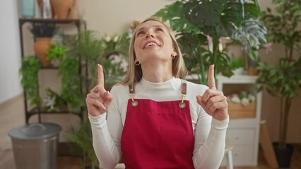 Poster - Stunned, young, attractive blonde florist sits joyfully at her flower shop table, amazed as she points up, looking surprised. a beautiful, cheerful portrait of positive adult happiness.