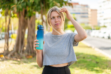  Young blonde sport woman with a bottle of water at outdoors having doubts and with confuse face expression