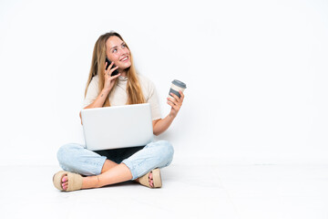 Young woman with laptop sitting on the floor isolated on white background holding coffee to take away and a mobile