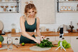Charming strong woman in green sport outfit cutting salad and recording video on mobile about healthy food. Fitness blogger with blond hair standing on kitchen and making tutorial